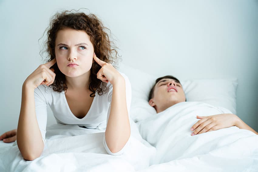 woman plugging her ears in bed while partner snores next to her