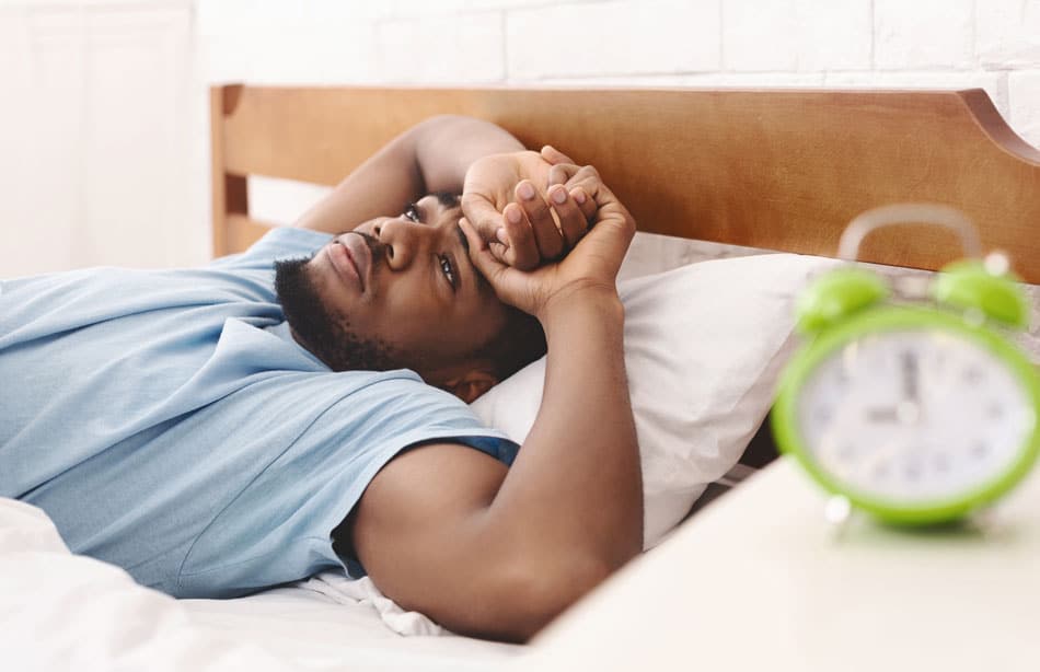 second-opinion-sleep-apnea-detroit Man lying awake in bed beside an alarm clock, illustrating insomnia, fatigue, or poor sleep quality.