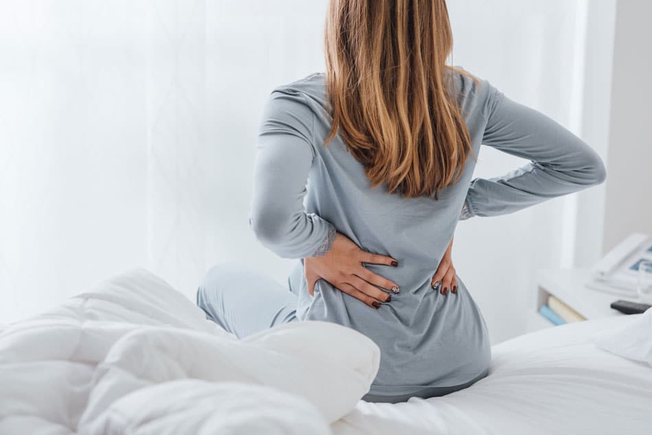 Woman sitting up in bed holding her lower back, illustrating morning back pain, muscle tension, or poor sleep posture.
