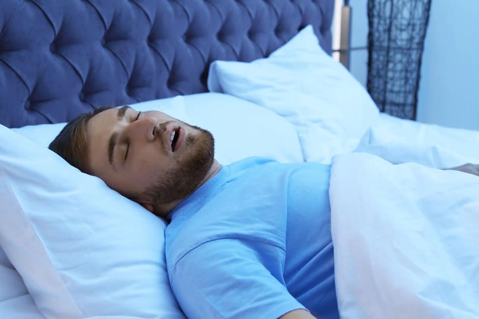 snoring-dangers-detroit Man sleeping on his back with mouth open, illustrating snoring or sleep apnea symptoms.