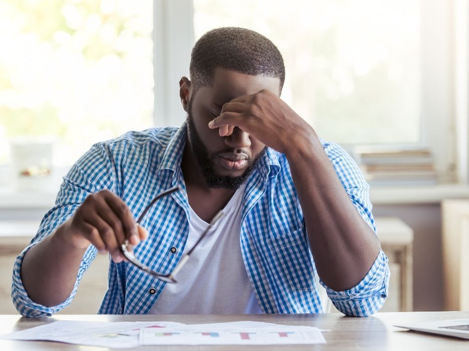 A man in a blue button-up sitting at a table with glasses in right hand using left hand to rub his forehead in discomfort