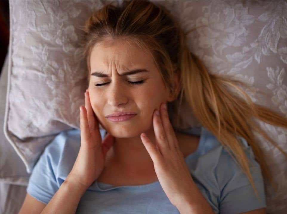 A woman laying in bed grabbing her jaw in discomfort