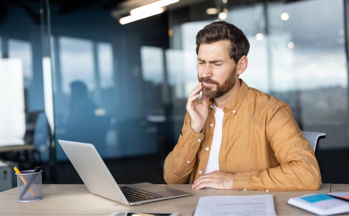 Man touching his jaw while working at a laptop, illustrating TMJ pain or dental discomfort affecting daily activities.